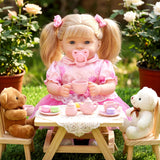 Blonde doll sitting at a wooden table with teacups, plush teddy bear, and white companion.