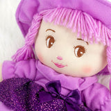 A purple hooded rag doll with yarn hair and stitched features rests near a large sparkly purple bow.