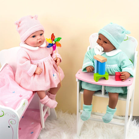A pink-dressed doll sits in a crib and a green-dressed doll sits at a chair.
