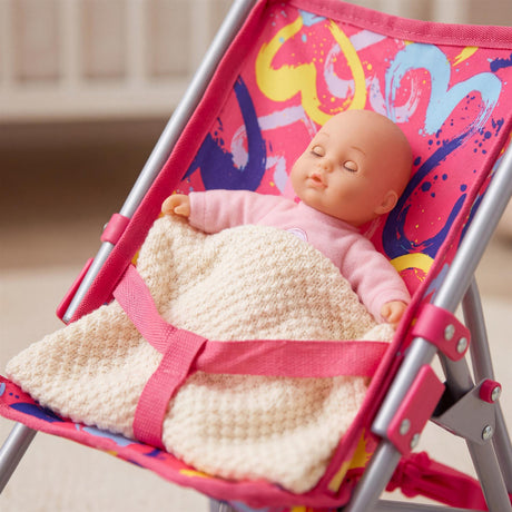 Doll rests in a pink patterned buggy, covered by a creamy knitted blanket and secured with a pink strap.
