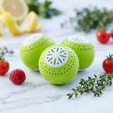 Three bright green, perforated balls are displayed with fresh herbs, raspberries, and tomatoes.