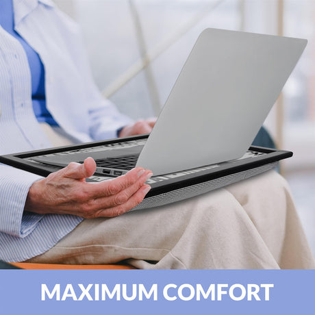 A person sits on a couch, holding a laptop on their lap, with a laptop stand underneath. The laptop is on a tray, and the person's hands are on the keyboard. The background is blurred, with a window visible in the distance. The person is wearing a blue shirt and a white jacket, and their hands are on the keyboard. The laptop stand is black and has a mesh design, and the laptop is a silver MacBook. - UKBuyZone