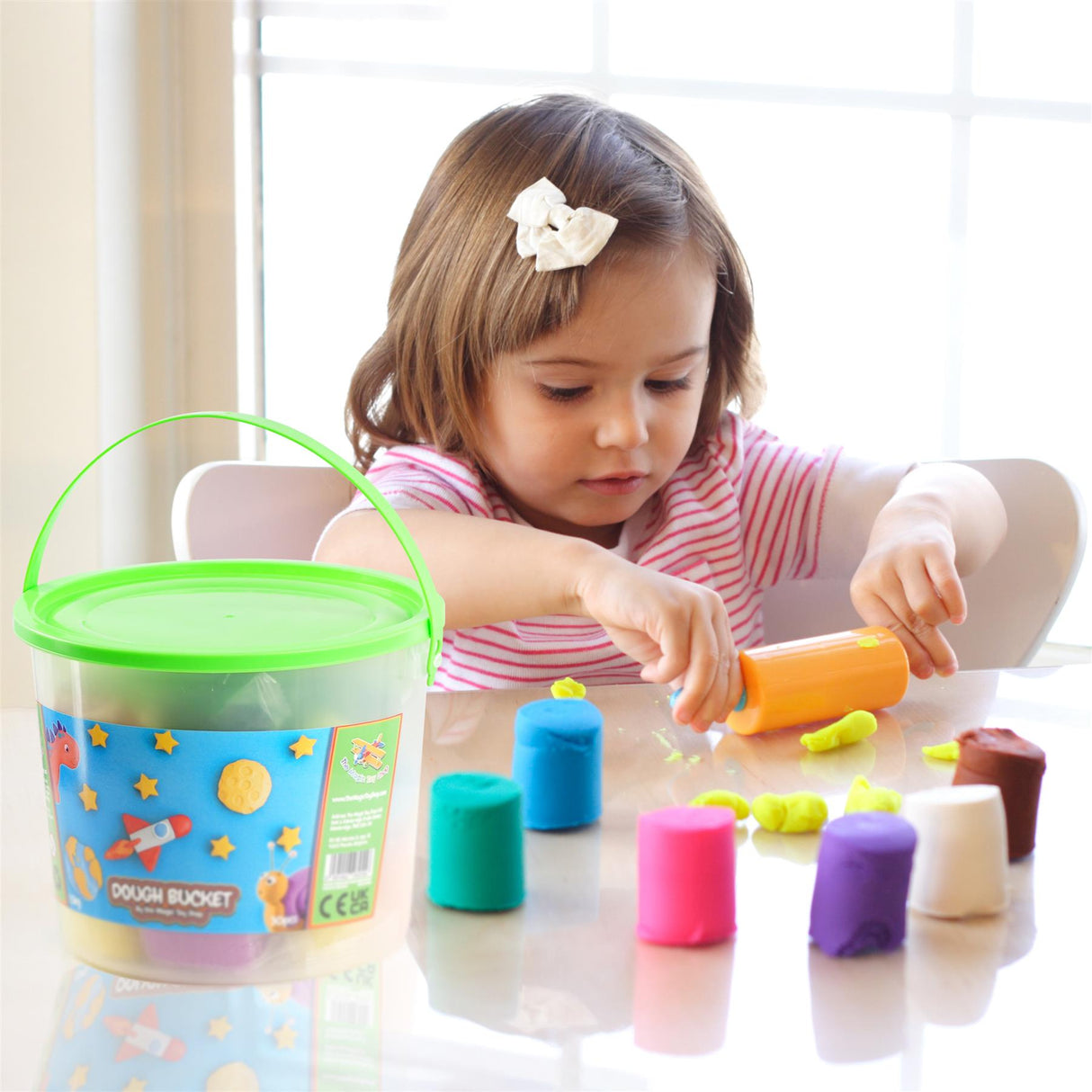 The image shows a young child playing with colored sand or clay. The child is using a small container to scoop the material into a bucket. There is a large plastic bucket on the table.