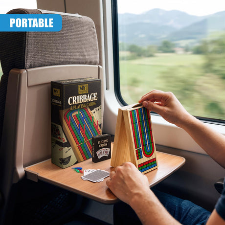 A person plays a wooden cribbage board game on a train tray table, next to its packaging and loose playing cards