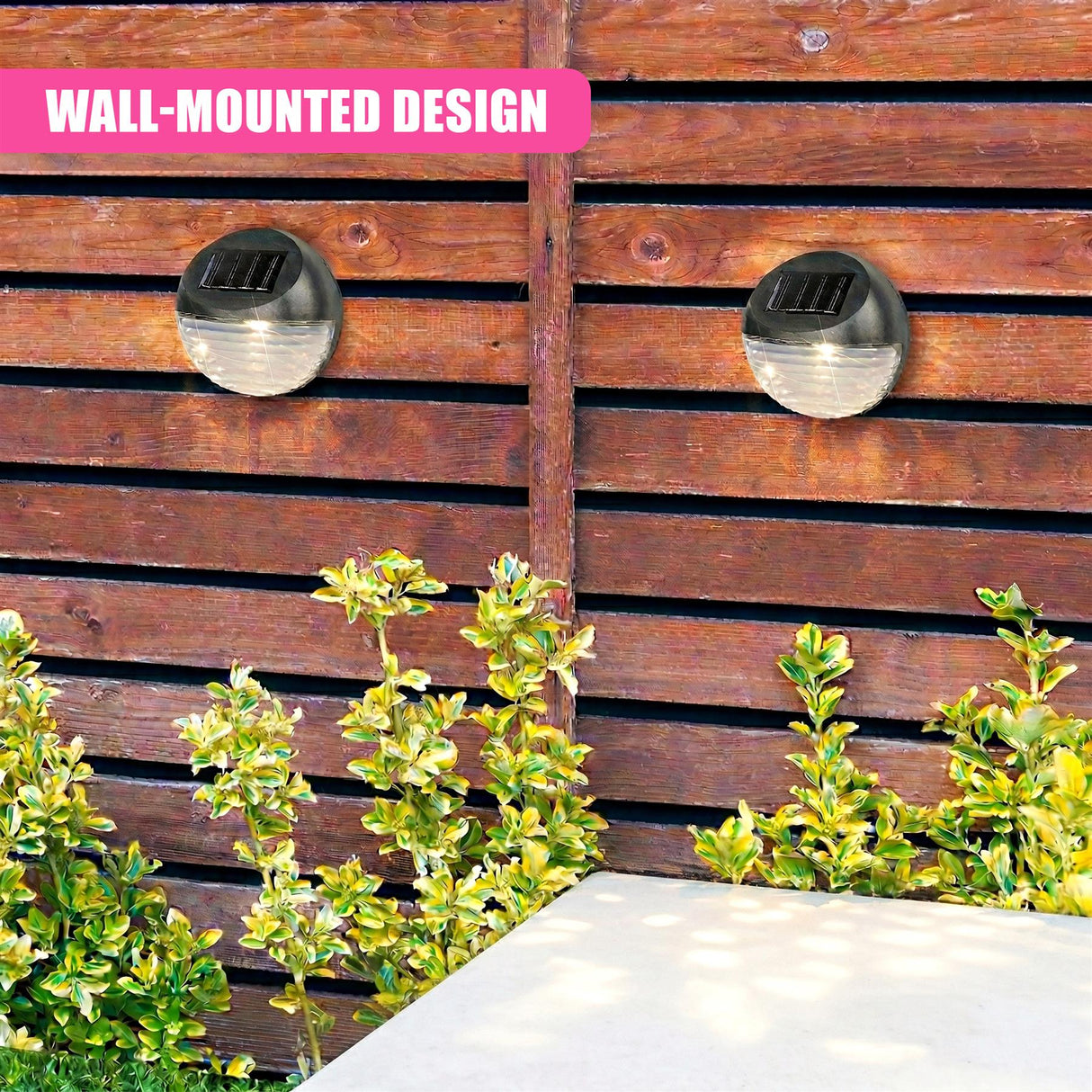 Two black, dome-shaped solar lights are mounted on a wooden fence next to green shrubs.