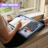 A woman sits on a window seat, surrounded by a tray of art supplies. She holds a sketchbook and pencil, while her feet are on a cushion. The tray is filled with a book, pencils, and a cup of coffee. A window is visible in the background, with a garden outside. The woman's face is not visible, but her hands and feet are. The scene is set in a peaceful, natural setting. - UKBuyZone