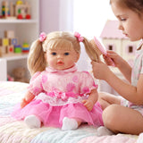 A girl brushes the hair of a blonde doll dressed in a pink and white outfit while sitting on a bed.
