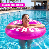 Smiling woman floating on a pink inflatable ring with white beach lettering in a blue pool.