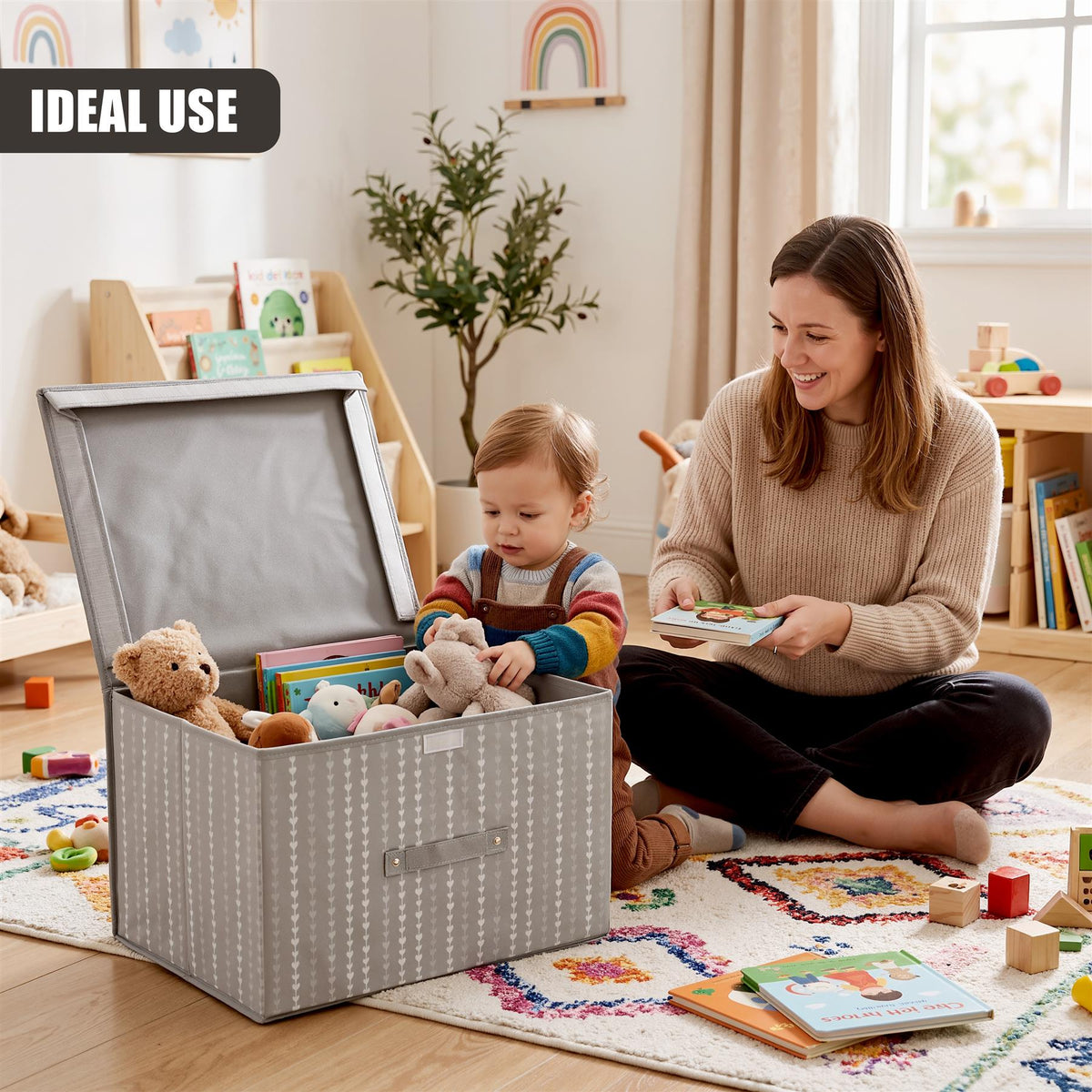 A toddler sits on a rug next to a large gray heart-patterned storage box filled with toys, while a woman reads a book nearby