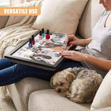 A woman sits on a couch, painting her nails while holding a tray with nail polish, a nail file, and a bowl of water. A small dog sleeps at her feet. The woman is surrounded by a cozy atmosphere, with a soft blanket and a few pillows. The tray has a fun design with a dog theme, featuring a dog's face and the phrase "I'd rather be with my dog." - UKBuyZone