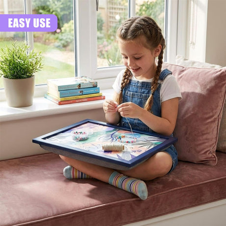 A young girl with braided hair sits on a couch, holding a tray with a colorful craft project. The tray has a blue frame and a design of leaves and flowers. The girl is surrounded by books and a potted plant, with a window in the background showing a garden. - UKBuyZone