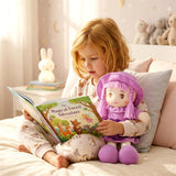 The rag doll wears a purple hat and dress while the girl reads a book together on the bed.