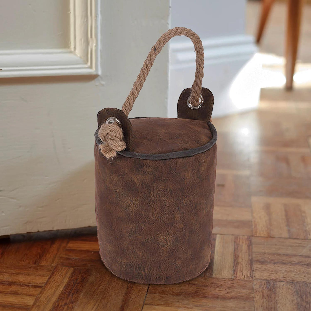 A brown faux leather bucket sits on a wooden floor, holding a rope. A handle is attached to one side of the bucket, and there's an embroidered flower detail on its surface. The bucket is placed near a wall, suggesting it might be serving as a door stopper. - UKBuyZone