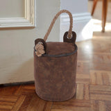 A brown faux leather bucket sits on a wooden floor, holding a rope. A handle is attached to one side of the bucket, and there's an embroidered flower detail on its surface. The bucket is placed near a wall, suggesting it might be serving as a door stopper. - UKBuyZone