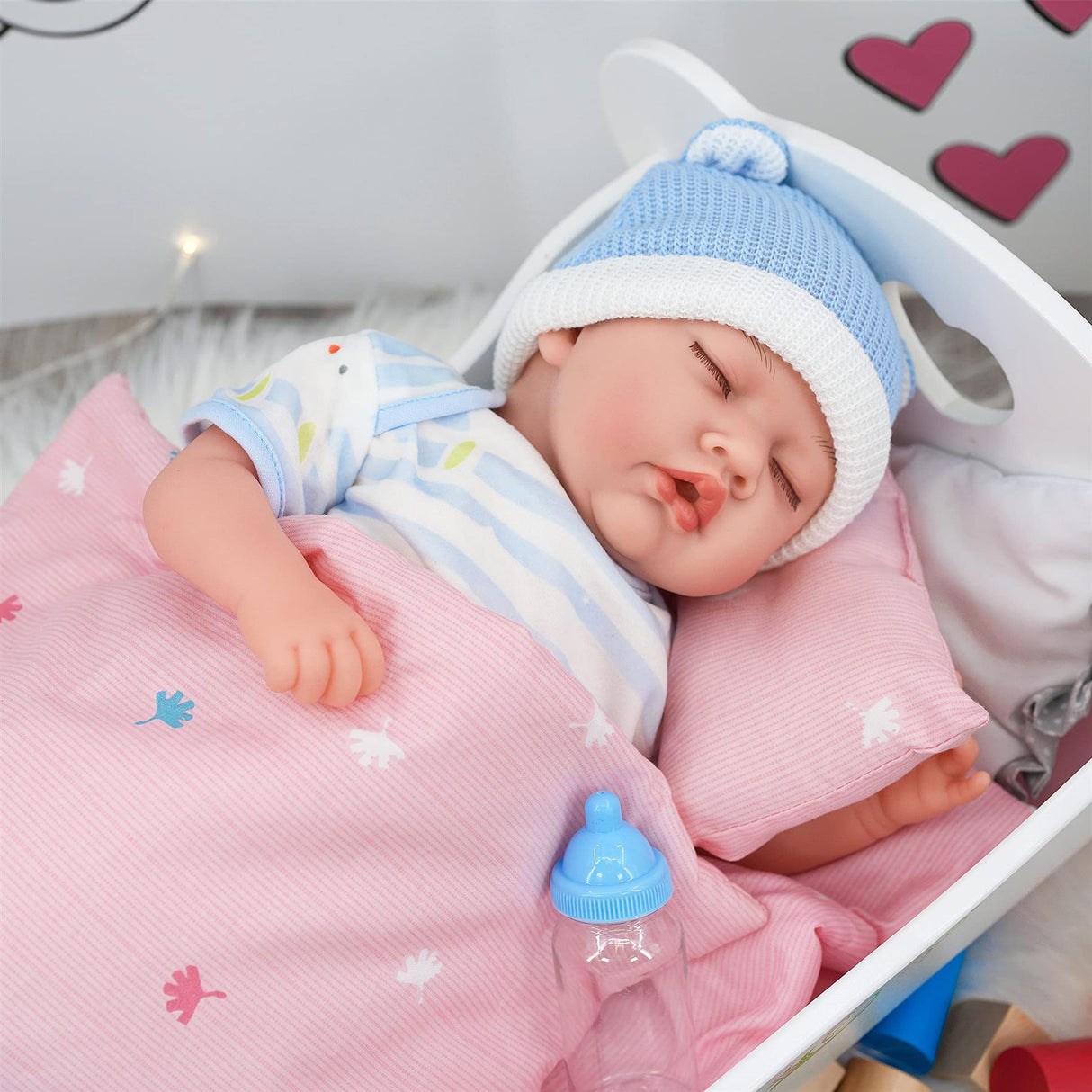A sleeping doll, wearing stripes and a blue hat, rests in a wooden cradle with a pacifier on the bed.