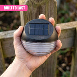 A hand holds a black, dome-shaped solar fence light fixture with visible photovoltaic panels.