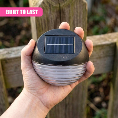 A hand holds a black, dome-shaped solar fence light fixture with visible photovoltaic panels.